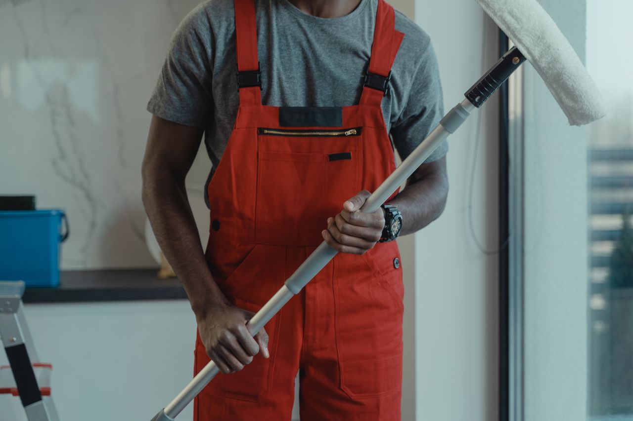 home-services-01 A professional cleaner wearing red overalls is holding a mop while cleaning indoors.